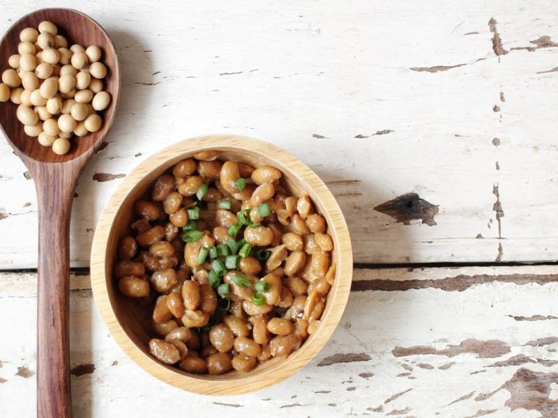 Natto in einer weißen Schüssel auf weißem Hintergrund. Links auf dem Foto ein Löffel mit Sojabohnen.