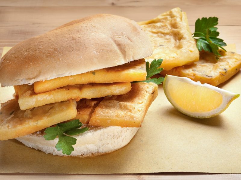 Panelle siciliane in einem aufgeschnittenen Brötchen. Daneben ein Zitronenschnitz.