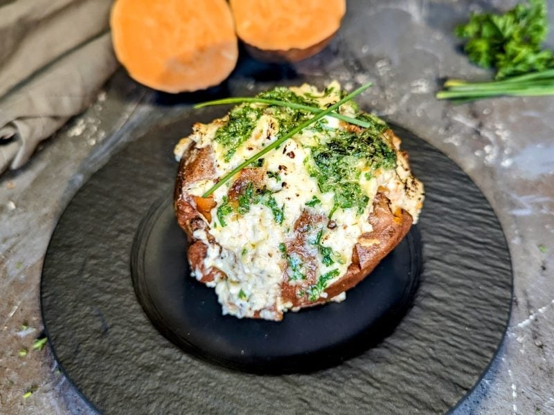 Ein schwarzer Teller mit der überbackenen Süßkartoffel mit Feta-Knoblauch-Creme auf grauem Steinuntergrund.