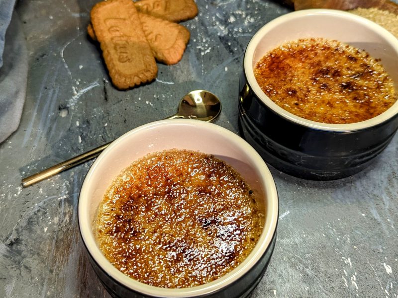 Zwei schwarze Schälchen mit der Biscoff Crème brûlée auf einem grauen Tisch mit den Keksen und grauem Tuch. Alles von schräg oben fotografiert.