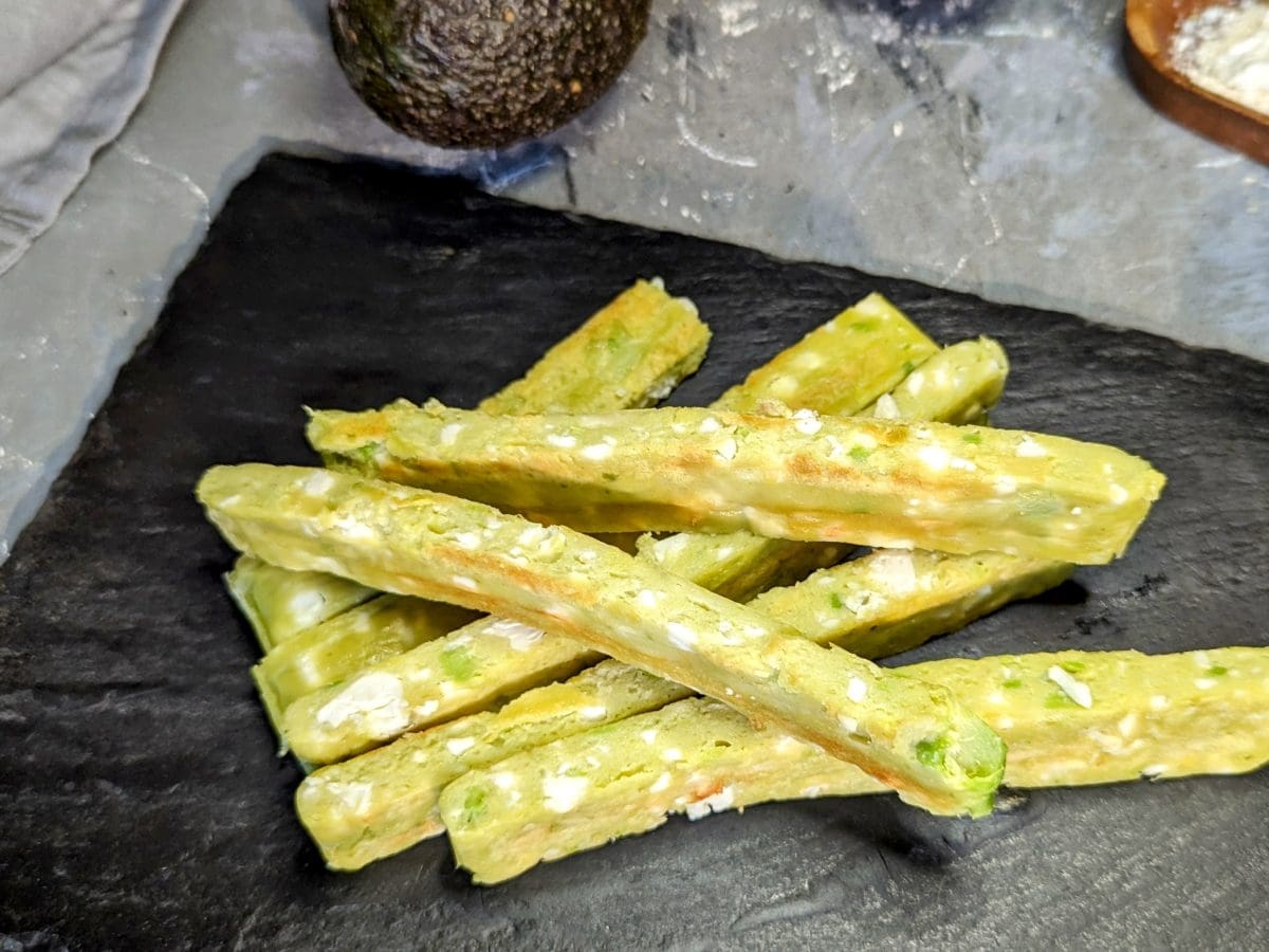 Mehrere Avocado-Feta-Stangen auf einer schwarzen Schieferplatte und grauem Steinuntergrund. Alles von schräg oben fotografiert.