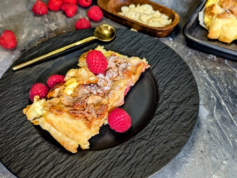 Eine Bienenstich-Schnitte mit Himbeeren auf einem schwarzen Teller mit goldenem kleinen Löffel auf grauem Steinuntergrund. Alles von schräg oben fotografiert.