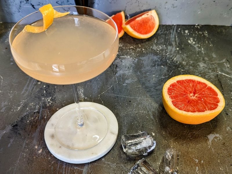 Eine Cocktailschale mit dem Drink auf Steinuntergrund von schräg oben fotografiert. Daneben Grapefruit und Cocktailzubehör.