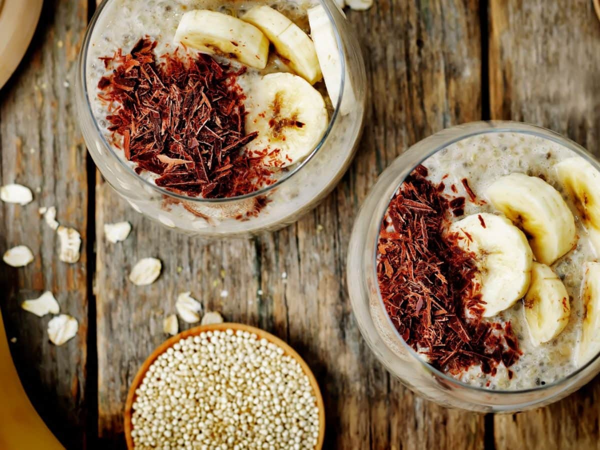 Zwei Gläser mit Quinoa-Pudding. Unten eine kleine Schüssel mit Quinoa und eine Banane. Der Hintergrund ist hölzern.