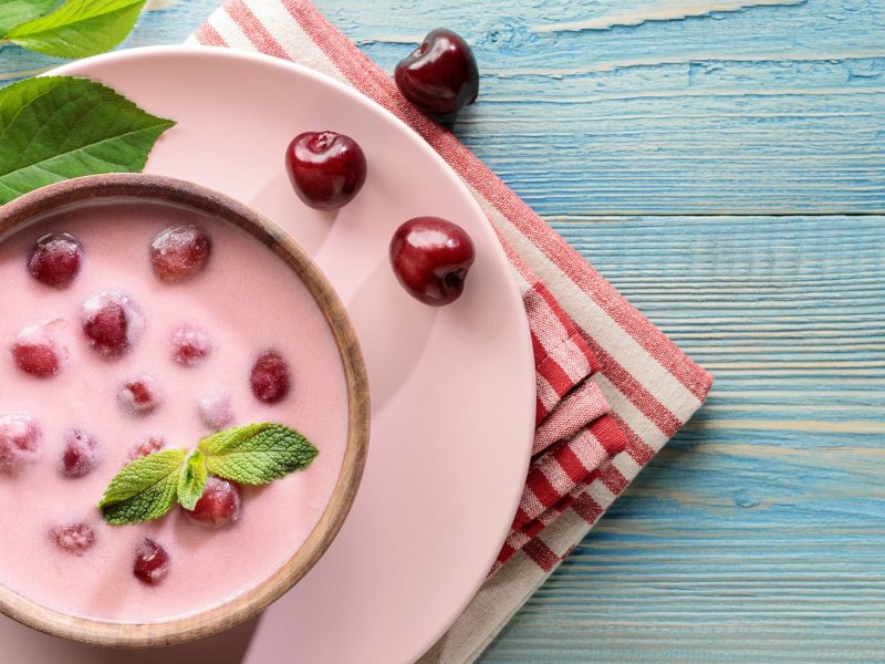 Ungarische Sauerkirschsuppe in einer Schüssel, die auf einem rosa Teller und Geschirrtuch steht. Daneben einzelne Kirschen.