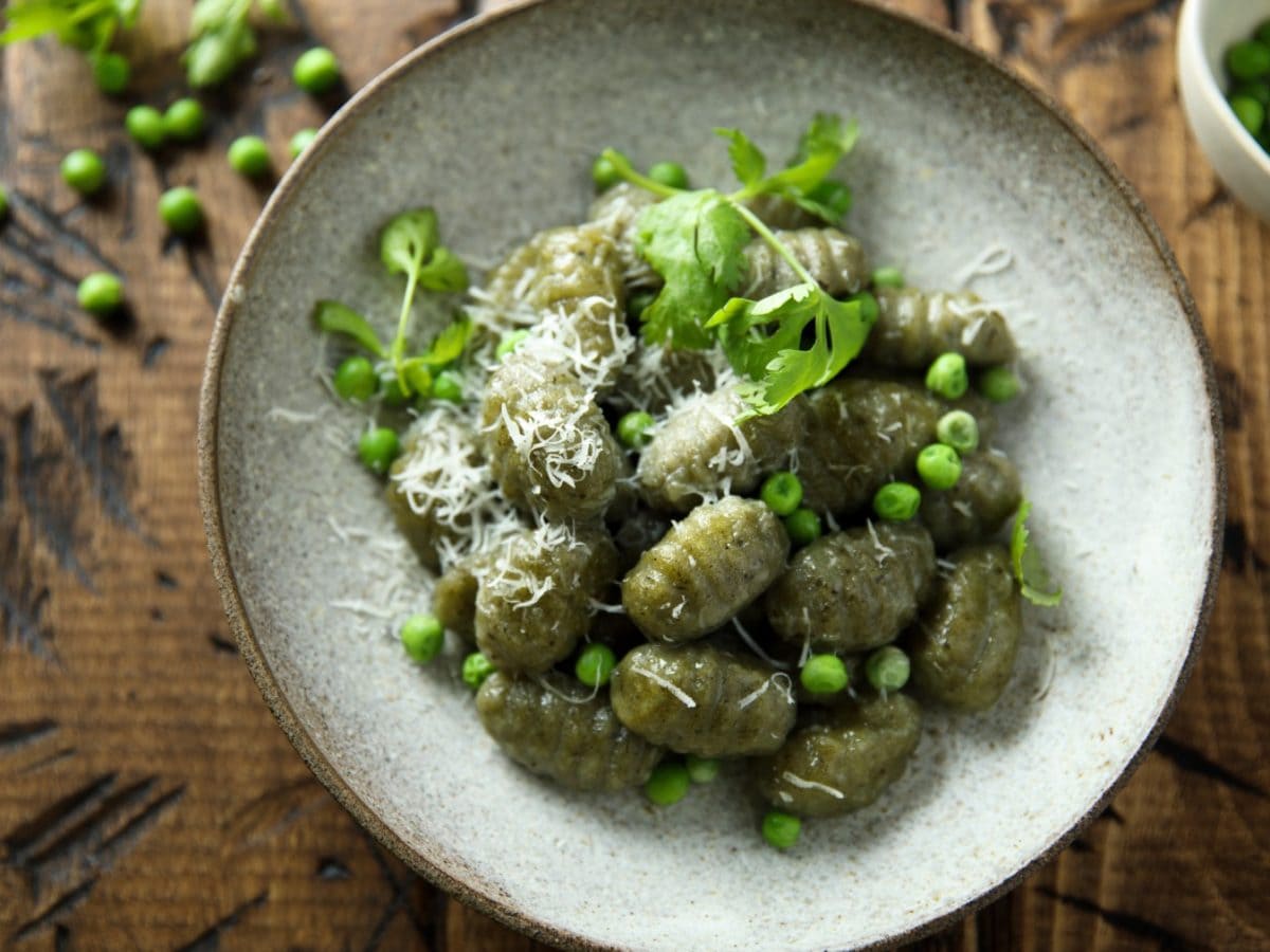 Spinat-Gnocchi auf einem grauen Teller auf hölzernem Hintergrund. Auf dem Hintergrund Erbsen verteilt und rechts eine kleine Schüssel mit Erbsen.