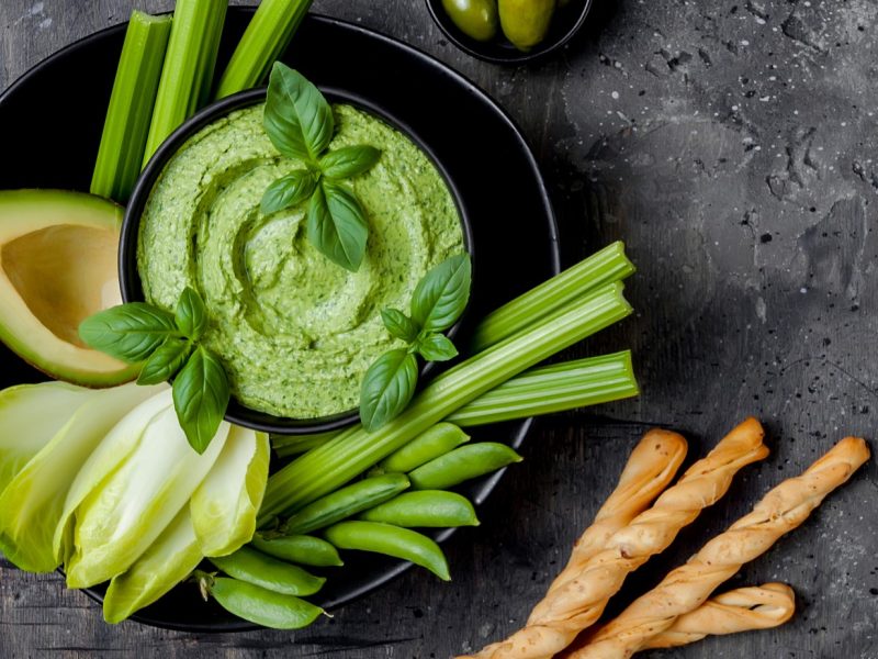 Ein schwarzer Teller mit dem Staudensellerie-Pesto und grünem Gemüse direkt von oben auf dunklem Untergrund fotografiert.