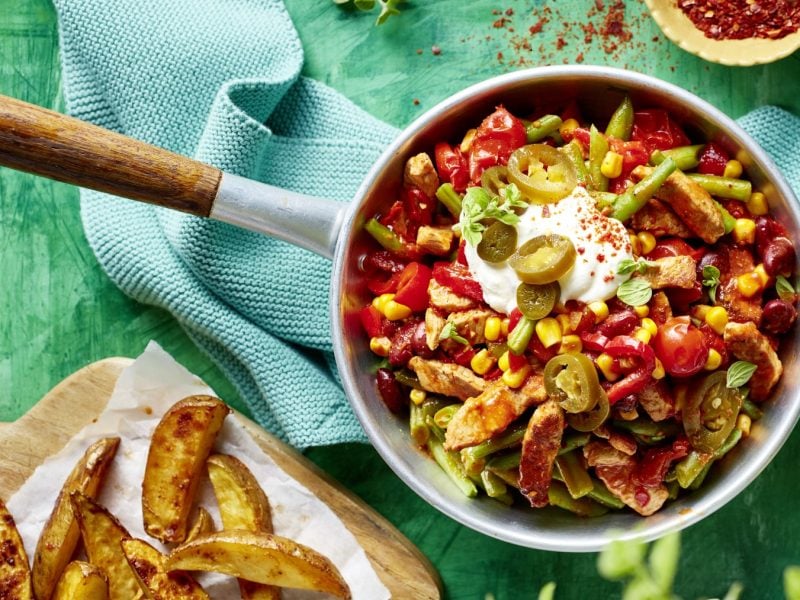 Tex-Mex-Pfanne vor einem türkisen Hintergrund auf einem blauen Geschirrtuch. Daneben ein Holzbrett mit Kartoffelspalten.