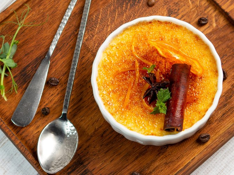 Thai-Tee Crème Brûlée in einer weißen Bowl auf einem braunen Schneidebrett. Links davon Besteck. Der Hintergrund ist hellgrau.