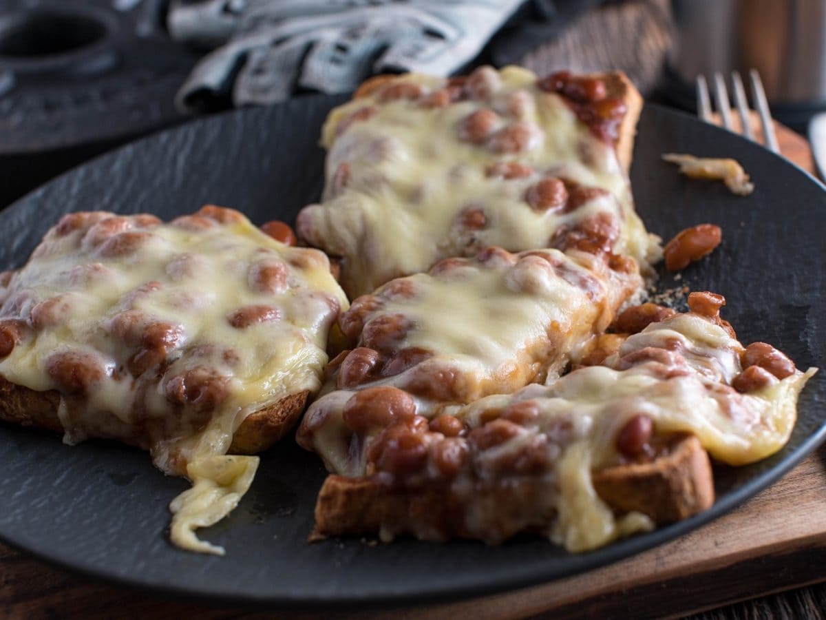 Draufsicht: Ein grauer Teller mit drei Broten. Es handelt sich um überbackener Baked-Beans-Toast, die mit Reibekäse bestreut sind.