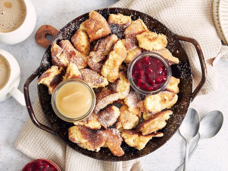 Veganer Kaiserschmarrn in einer schwarzen Pfanne mit Preiselbeermarmelade und Apfelmus serviert. Die Pfanne steht auf einem beigen Geschirrtuch und der Hintergrund ist hellgrau. Links auf dem Bild zwei Kaffeetassen, rechts zwei Löffel.