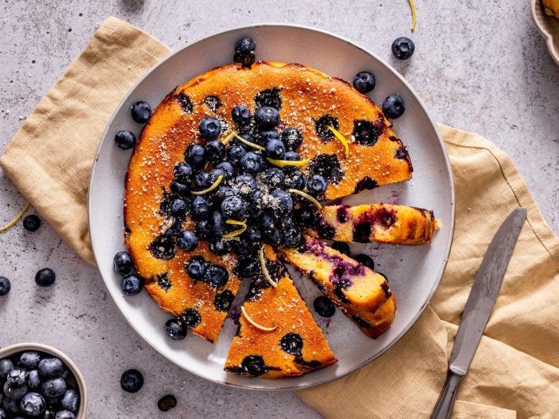 Draufsicht: Angeschnittener Veganer Limoncello-Kuchen mit Blaubeeren auf weißem Teller mit hellem Untergrund, drum herum Blaubeeren.