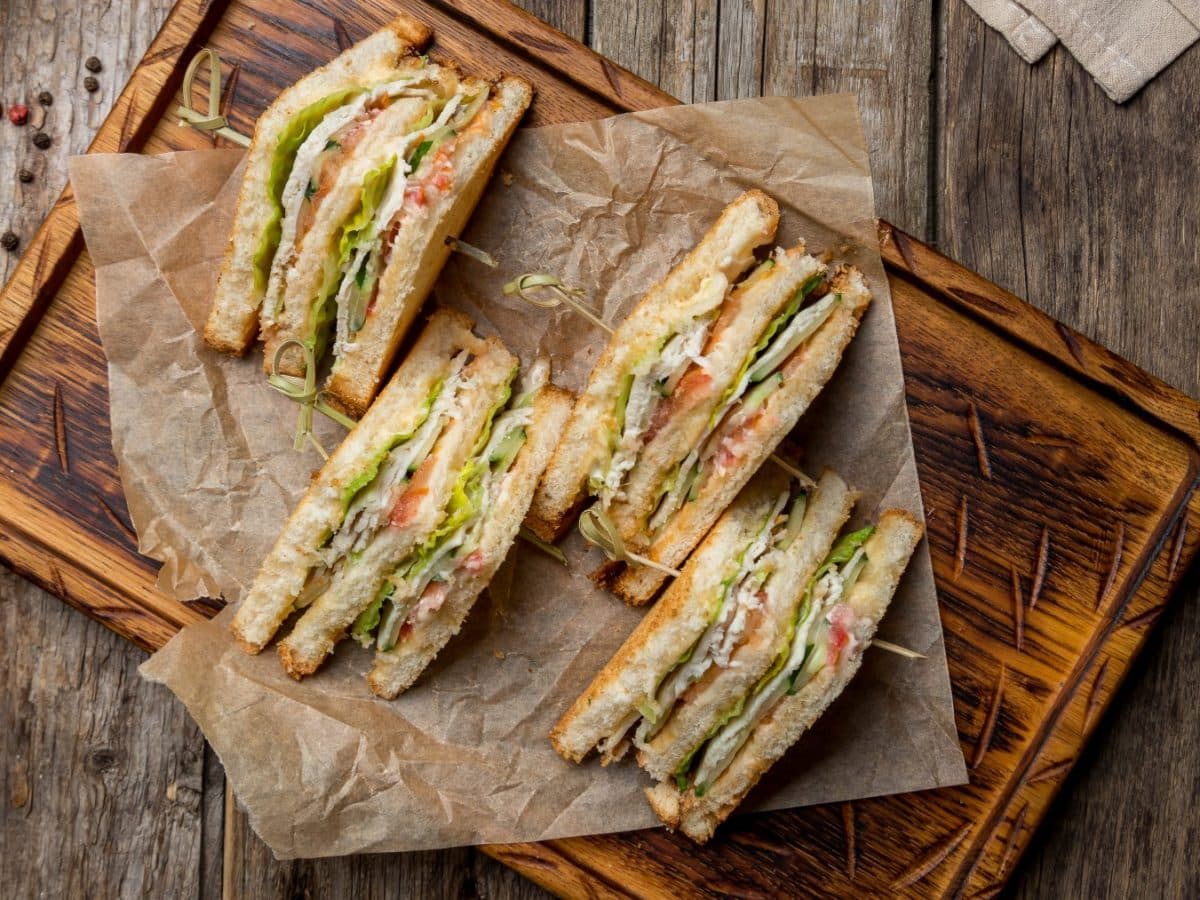 Veganes Club-Sandwich auf Backpapier auf einem Schneidebrett. Der Hintergrund ist hölzern und Pfefferkörner darauf verteilt.