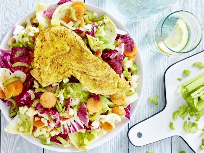 Wintersalat mit Putenschnitzel auf einem weißen Teller. Rechts davon ein Schneidebrett mit Staudensellerie und rechts oben auf dem Bild ein Glas Wasser mit Zitrone.