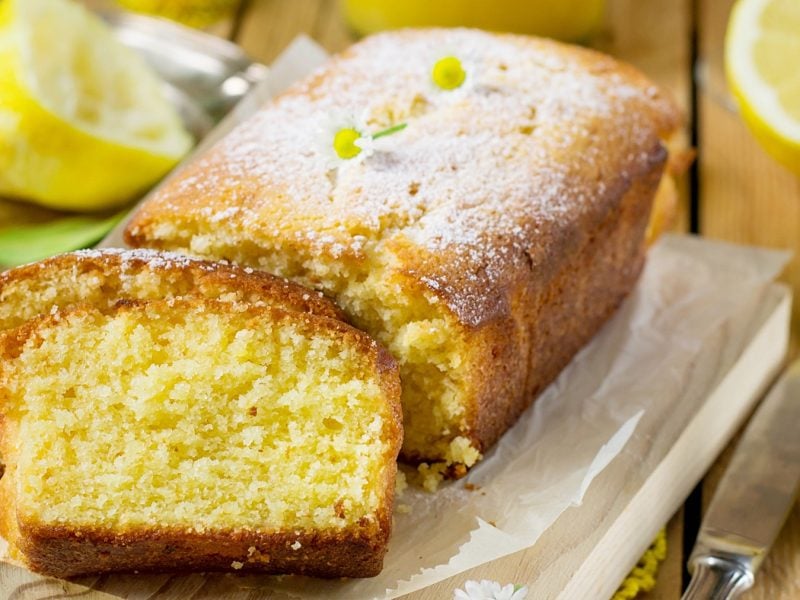 Der angeschnittene Zitronen-Kastenkuchen auf einem Holzbrett und rustikalem Holztisch. Daneben und dahinter gelbe Blüten und Zitronen. Alles schräg von oben und leicht von der Seite fotografiert.