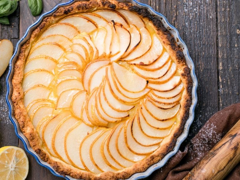 Draufsicht: Runde Zitronen-Tarte mit Birne auf Holzunterhgrund, drum herum Zitronen, Birnen und ein Nudelholz.