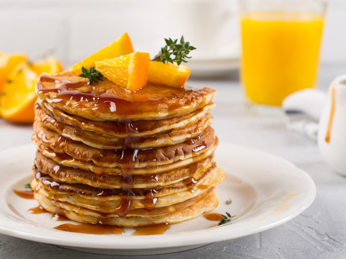 Zitronen-Pancakes mit Orangensirup auf einen Teller gestapelt mit Orangenstücken darauf und daneben, heller Hintergrund.