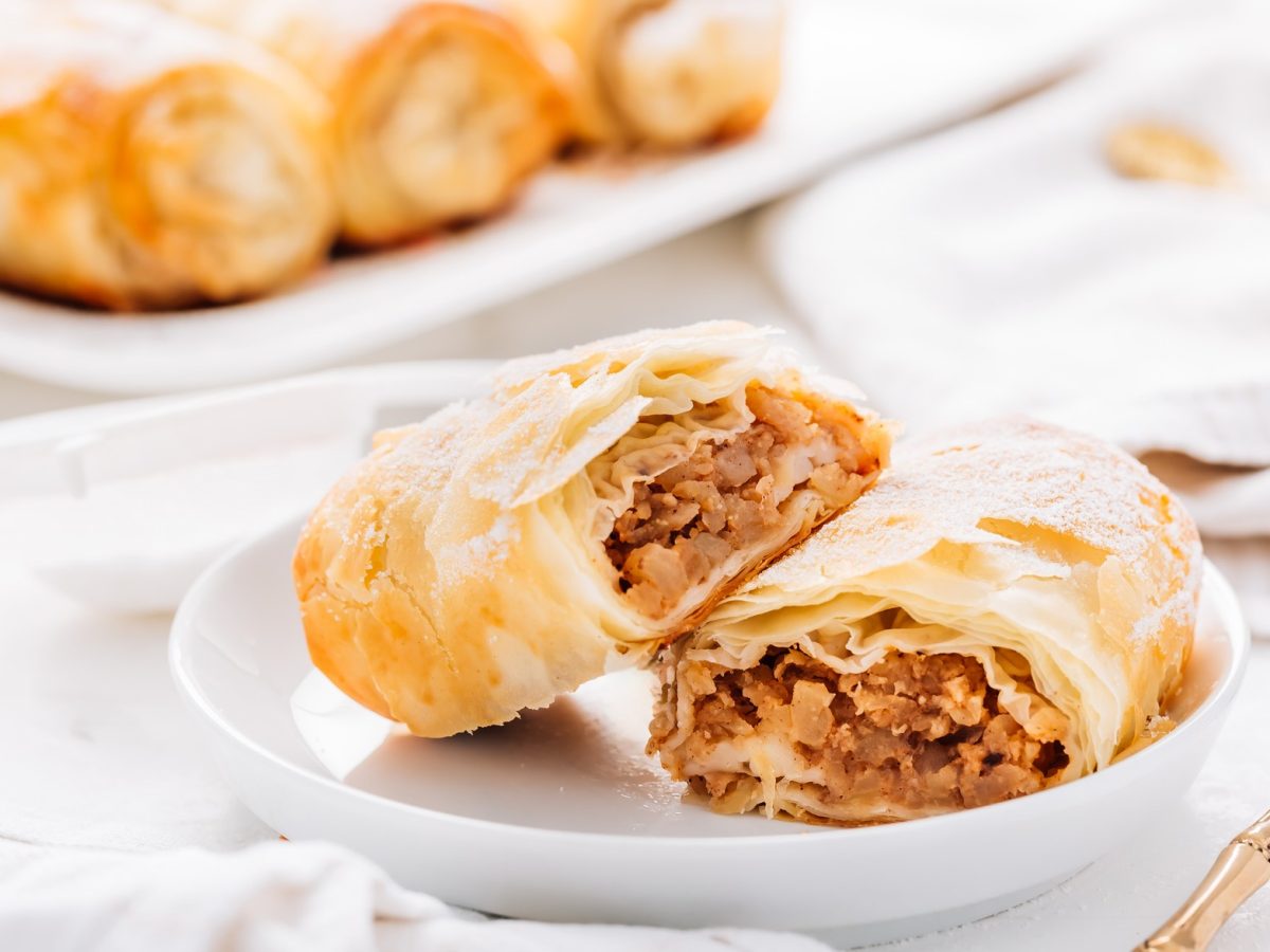 Ein weißer Teller mit einem halbierten Apfelstrudel-Röllchen und drei Stück im Hintergrund. Alles auf weißem Tisch mit weißer Tischdecke und sehr nah von der Seite fotografiert.