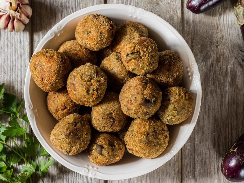 Draufsicht auf polpette di melanzane in einer weißen Schüssel. Drumherum Auberginen, Petersilie und Knoblauch.