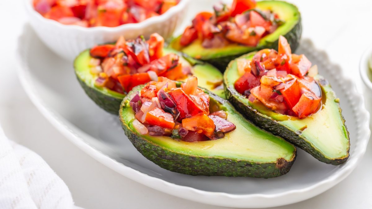 Vier ausgehöhlte Avocadohälften mit dem Tomatenragout auf hellem Teller und hellem Untergrund.