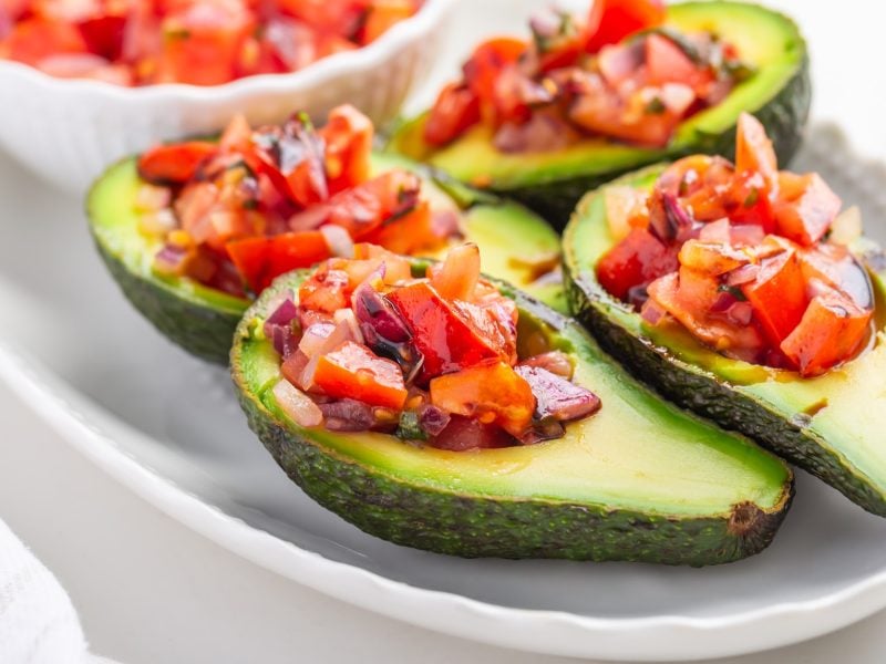 Vier ausgehöhlte Avocadohälften mit dem Tomatenragout auf hellem Teller und hellem Untergrund.