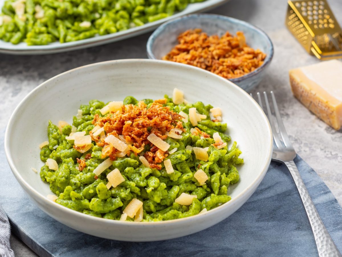 Ein weißer Teller mit den grünen Spätzle auf einem hellblauem Marmortisch mit grauem Tuch. Alles von schräg oben fotografiert.