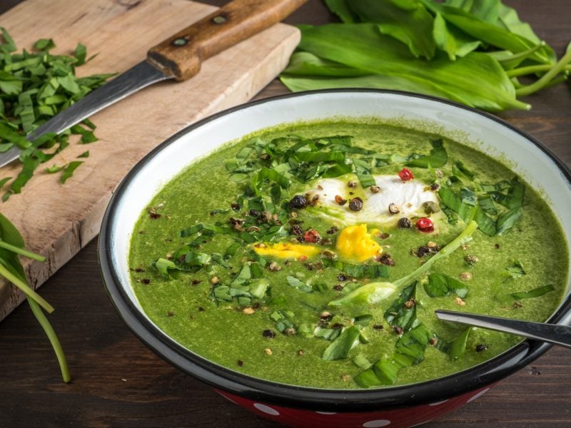 Die Bärlauchsuppe in einer hellen Schale auf einem Holztisch mit frischem Bärlauch im Hintergrund. Alles leicht oben von der Seite fotografiert.