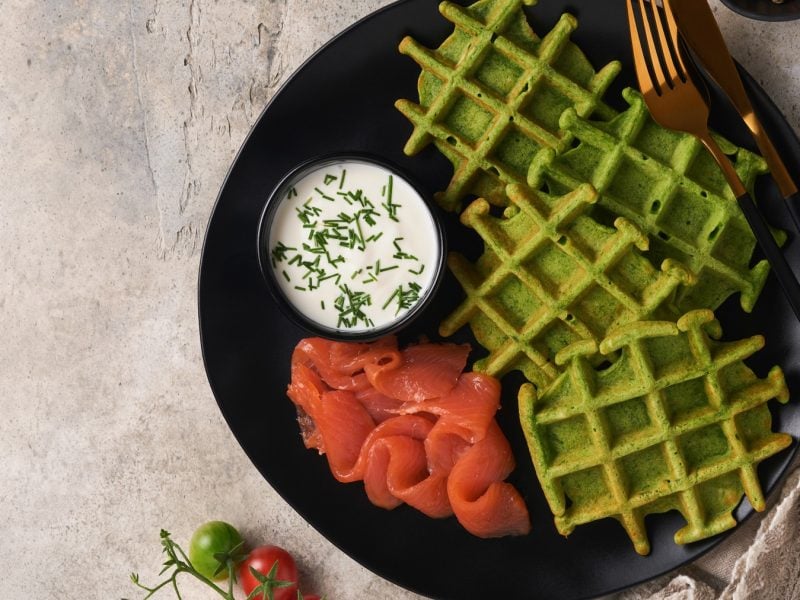 Ein schwarzer Teller mit den Bärlauchwaffeln und Räucherlachs auf grauem Steintisch. Alles direkt von oben fotografiert.