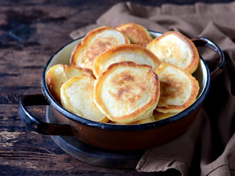 Ein kleiner brauner Topf mit den Bananenscheiben-Pancakes auf einem dunklen Holztisch. Alles von schräg oben fotografiert.