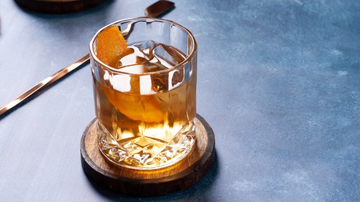 Zum Tag des Bourbon: Was ist Bourbon? Ein Glas mit Bourbon auf einem dunkelblauen Tisch mit Barlöffel. Frontalansicht.