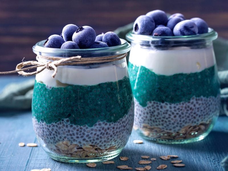 Zwei Gläser mit dem Chia-pudding auf einem blauem Tishc udn dunklem Holzhintergrund. Alles von der Seite fotografiert.