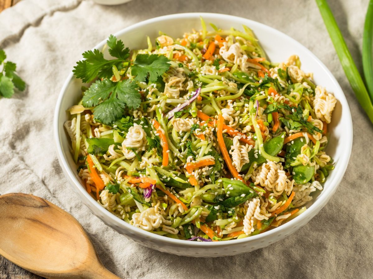 Eine weiße Schale mit dem crunchy Nudelsalat auf einem Holztisch mit cremefarbener Tischdecke. Alles von oben fotografiert.