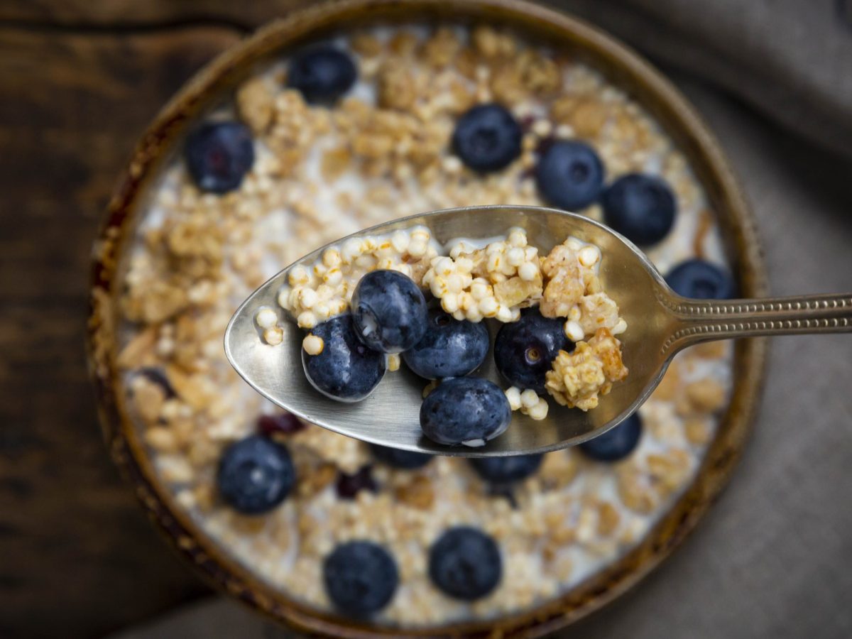 Draufsicht: Eine runde Beeren-Quinoa-Bowl steht auf einem Holztisch. Über der Bowl ist ein gefüllter Silberlöffel mit Beeren und Quinoa.