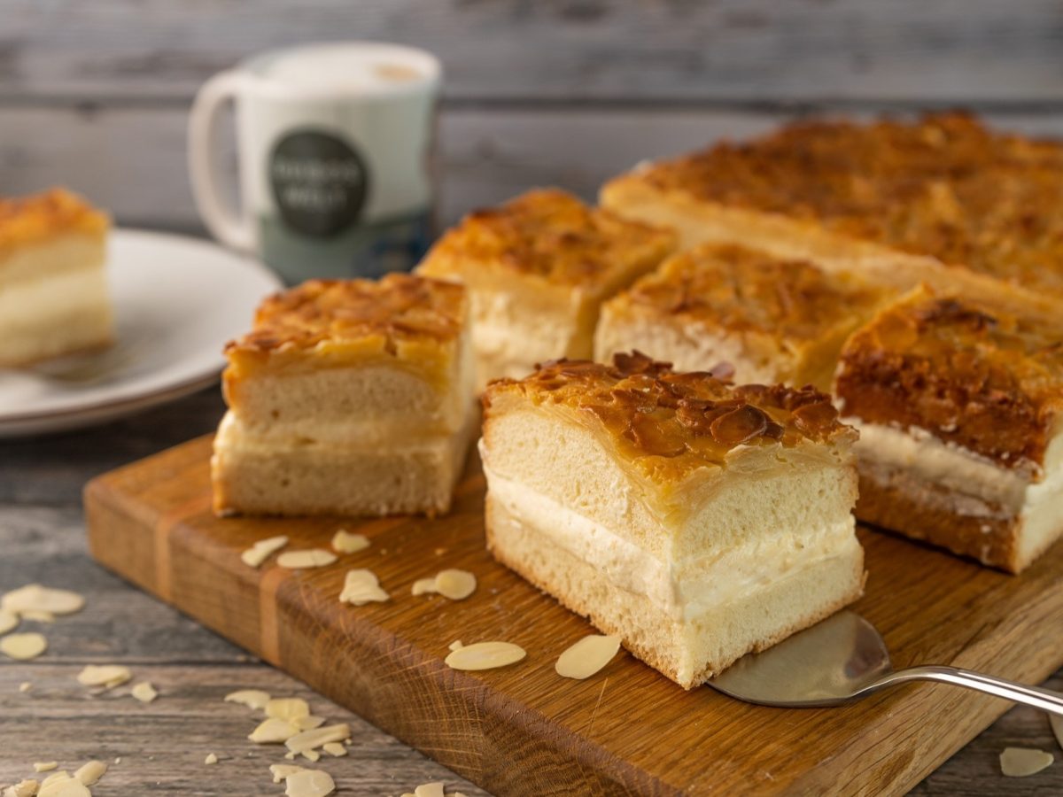 Frontal, leicht schräg von oben: Auf einem Holztisch liegt ein Holzbrett auf dem mehrere Stücke Bienenstich-Kuchen liegen. Im Hintergrund ist ein Teller mit einer Gabel und einem Stückchen Kuchen.