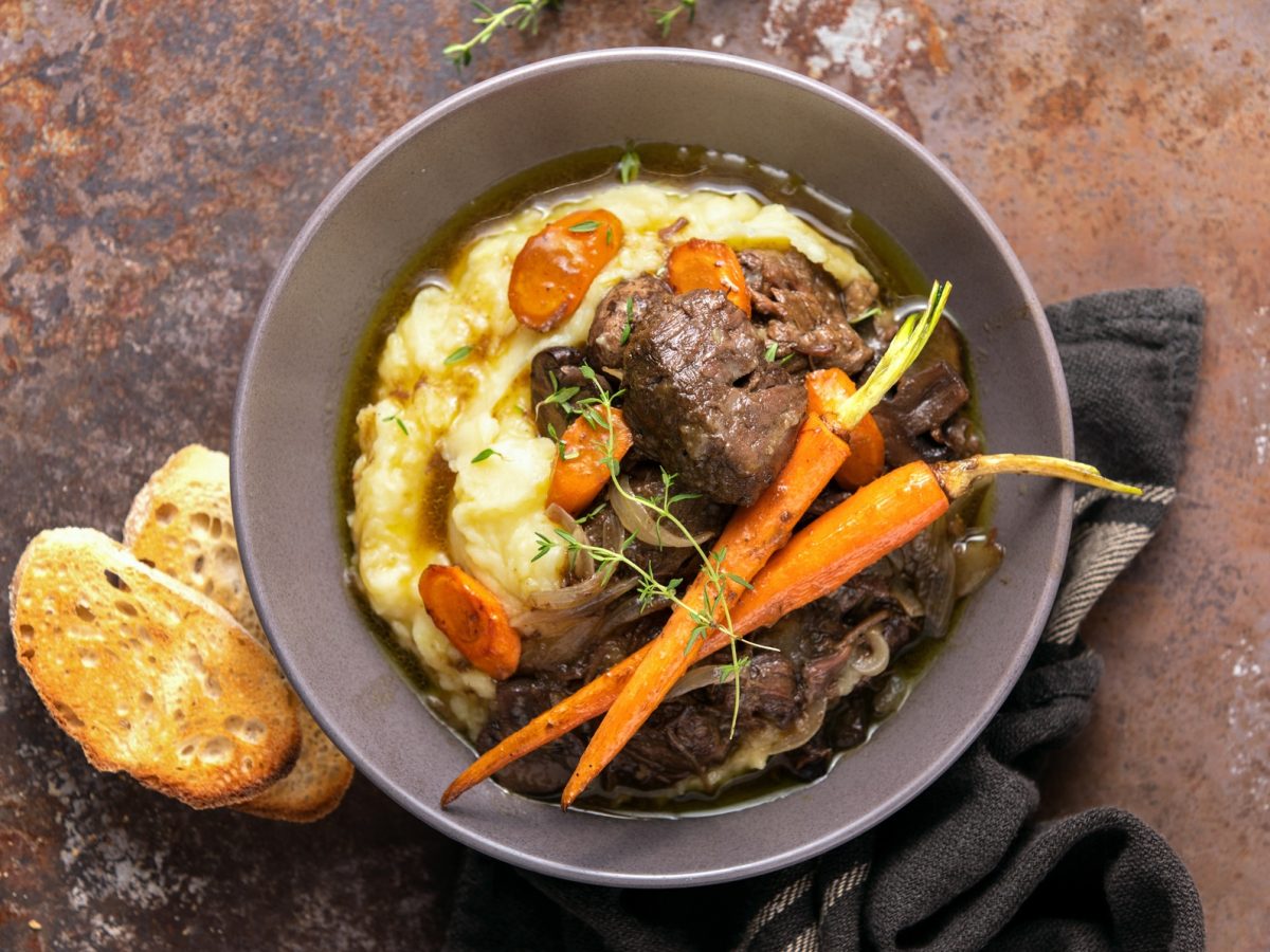 Draufsicht: Eine Ess-Schüssel Boeuf Bourguignon mit Kartoffelpüree steht auf einem rustikalen Untergrund aus Holz. Daneben liegt eine Scheibe geröstetes Weißbrot.