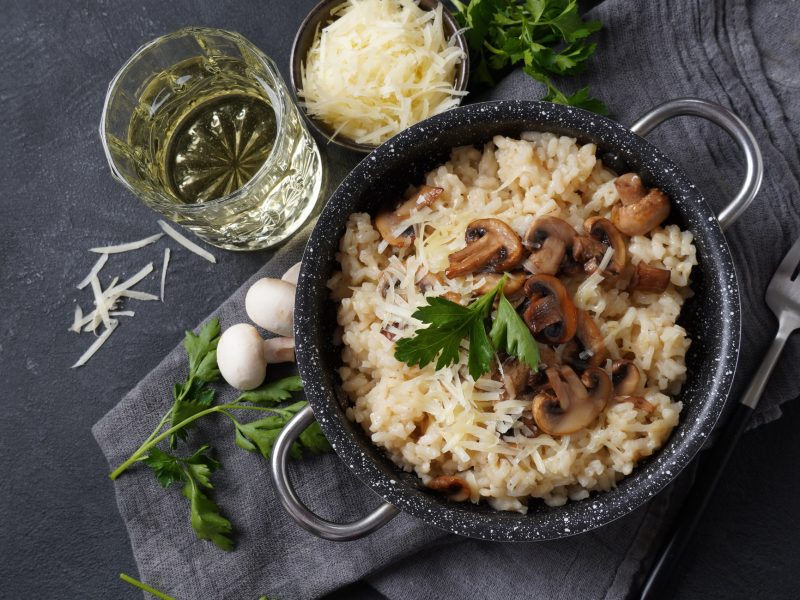 Draufsicht: Ein dunkler Topf Champignon-Risotto auf einem gräulichen Untergrund. Daneben sind Glas Weißwein, frische Pilze, frisch geriebener Käse und Petersilie.