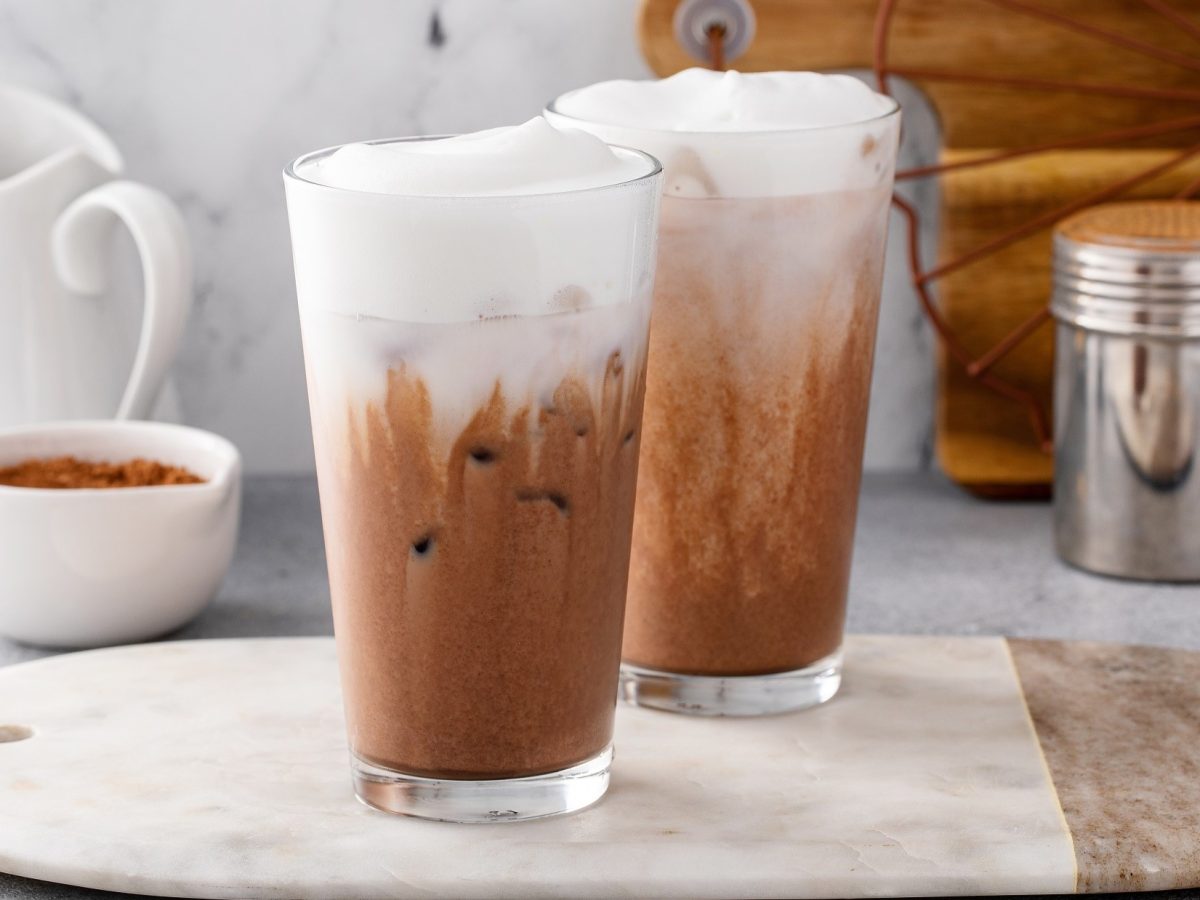 Frontal: Zwei Gläser Chocolate-Chai stehen auf einem Holzbrettchen. Das liegt auf einem weißen Untergrund aus Marmor. Naneben stehen eine weiße Milchkanne aus Porzellan, eine Tasse mit braunem Zucker und ein Kakao-Streuer aus Edelstahl.