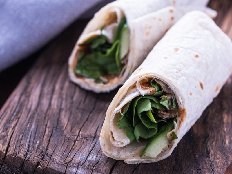 Draufsicht, schräg: Zwei gerollte Dürüm Çiğ Köfte liegen auf einem dicken und schmalen Holzbrett. Aus den Wraps lugen Salatblätter heraus.