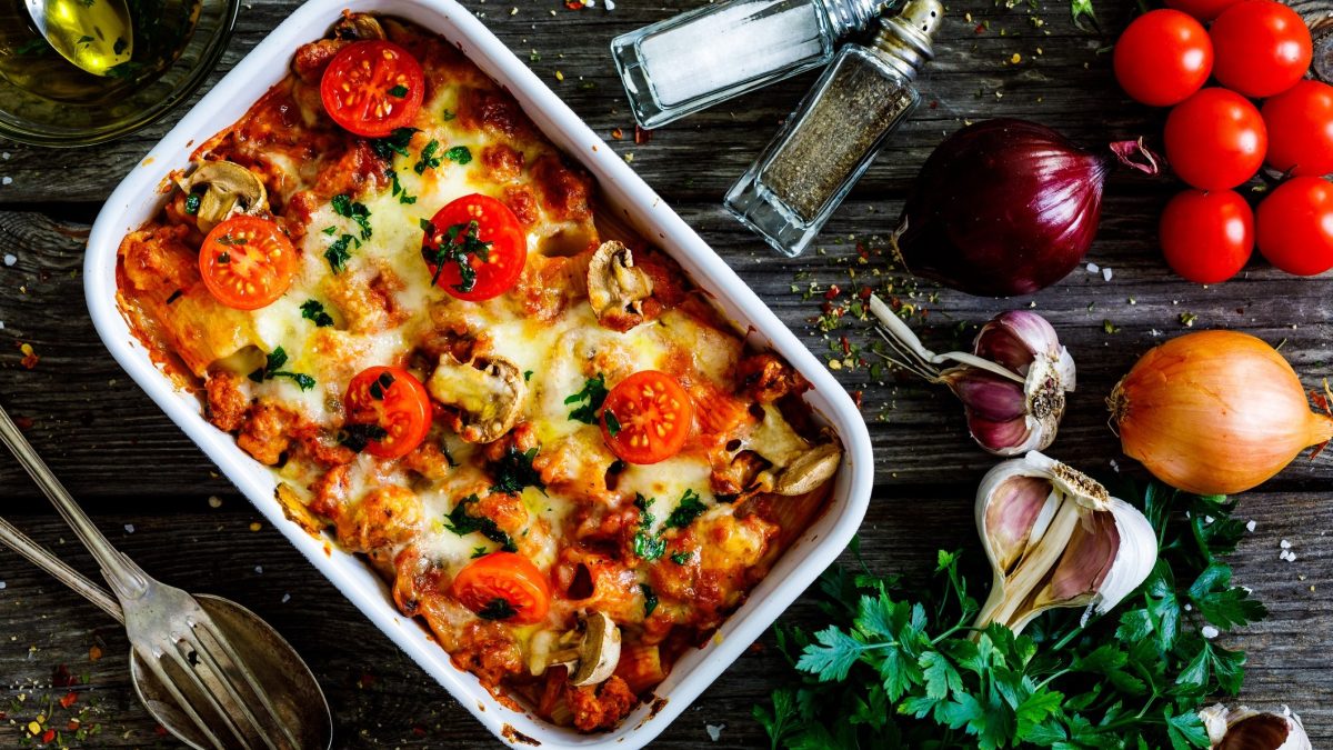 Draufsicht: Der Auslauf Faschiertes mit Pasta und Tomaten steht auf einem dunklen Untergrund aus Holz. Drum herum liegen Kräuter, sowie frische Zutaten, wie etwa Tomaten und Zwiebeln.