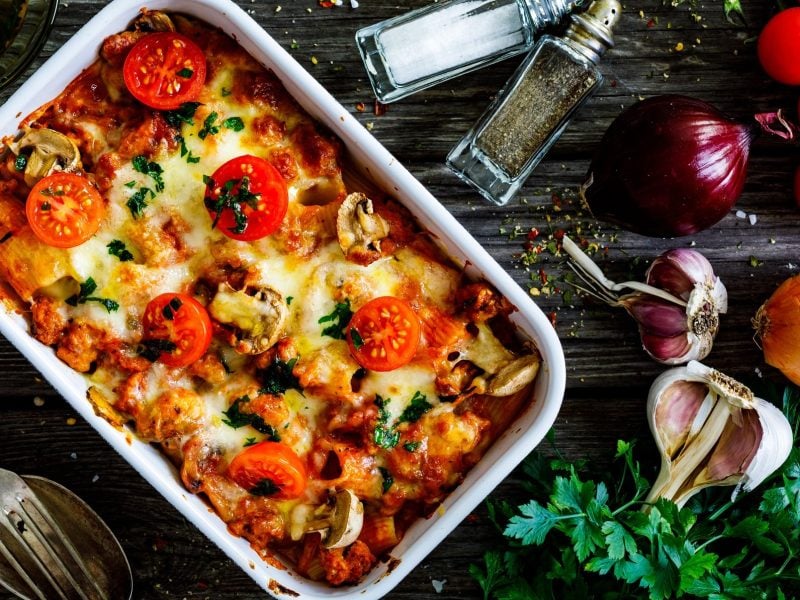 Draufsicht: Der Auslauf Faschiertes mit Pasta und Tomaten steht auf einem dunklen Untergrund aus Holz. Drum herum liegen Kräuter, sowie frische Zutaten, wie etwa Tomaten und Zwiebeln.