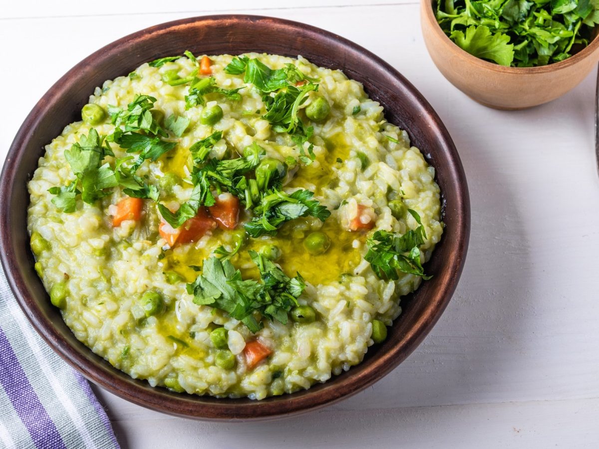 Draufsicht: Ein runder Teller Frühlings-Risotto mit Gartengemüse auf einem weißen Untergrund. Daneben liegt ein blau-weiß-gestreiftes Küchentuch. Darüber hinaus gibt es eine kleine Schale mit frischer Petersilie sowie eine Gabel.