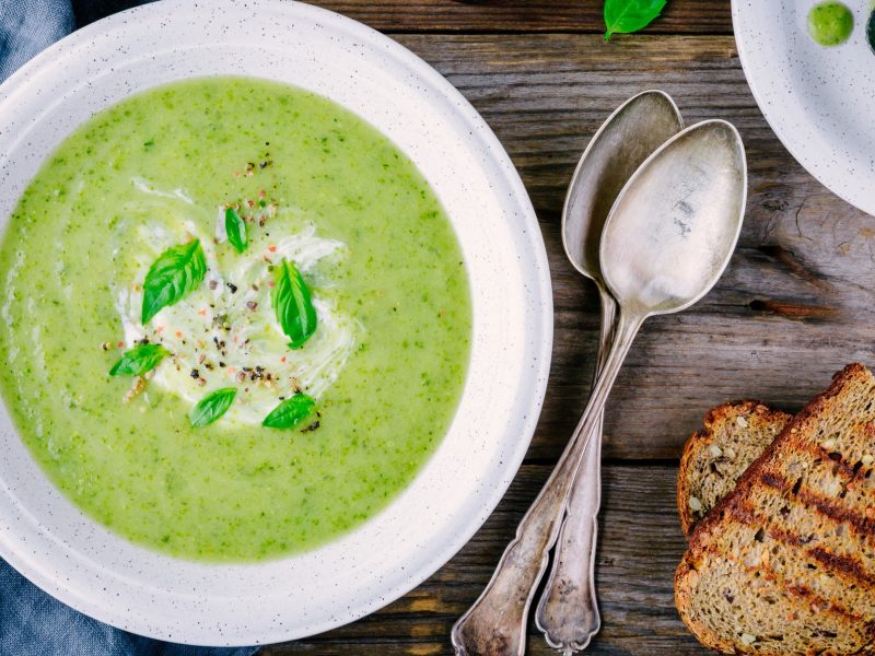 Draufsicht: ein weißer Teller mit einer Frühlingskräuter-Suppe mit Spinat steht auf einem Tisch. Daneben liegen zwei geröstete Brotscheiben und zwei Löffel.