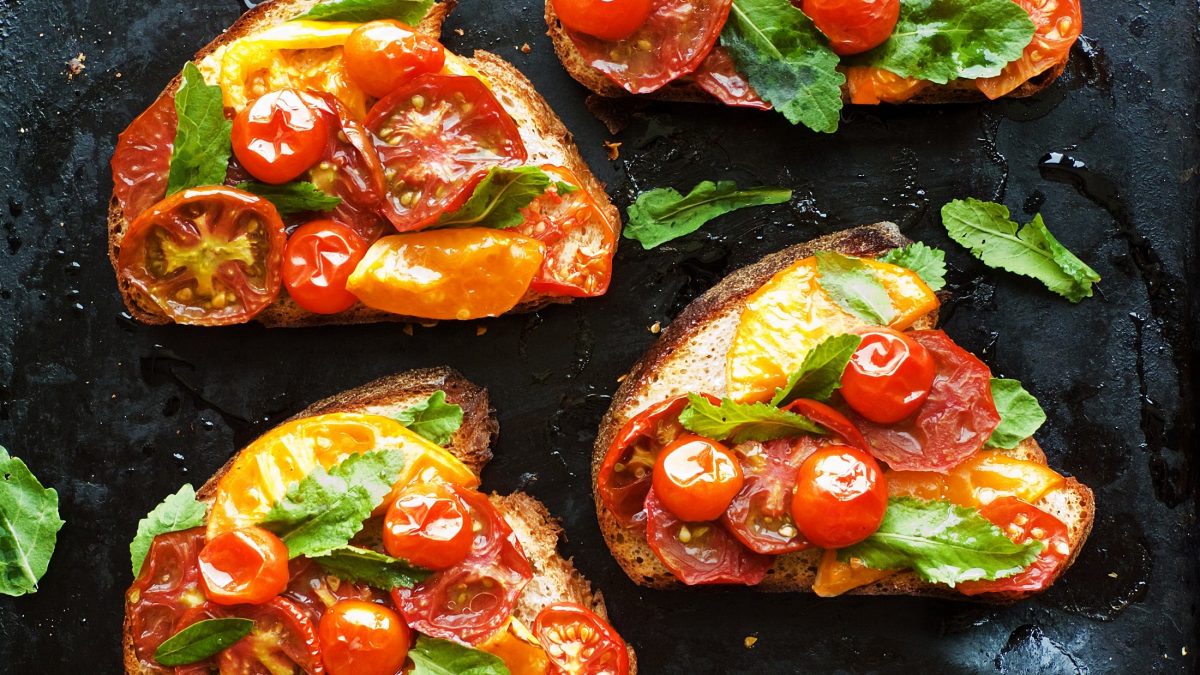 Draufsicht: Mehrere geröstete Tomatenbrote mit frischen Kräutern liegen auf einem schwarzem Untergrund.