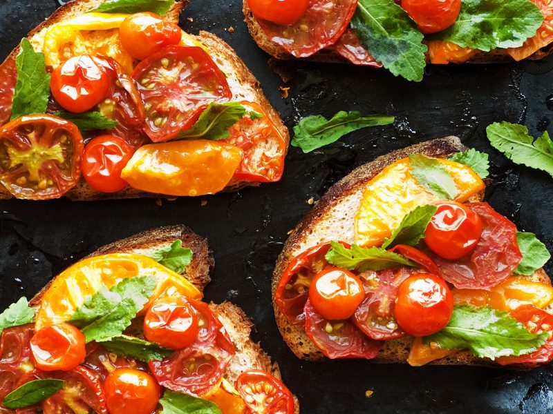 Draufsicht: Mehrere geröstete Tomatenbrote mit frischen Kräutern liegen auf einem schwarzem Untergrund.