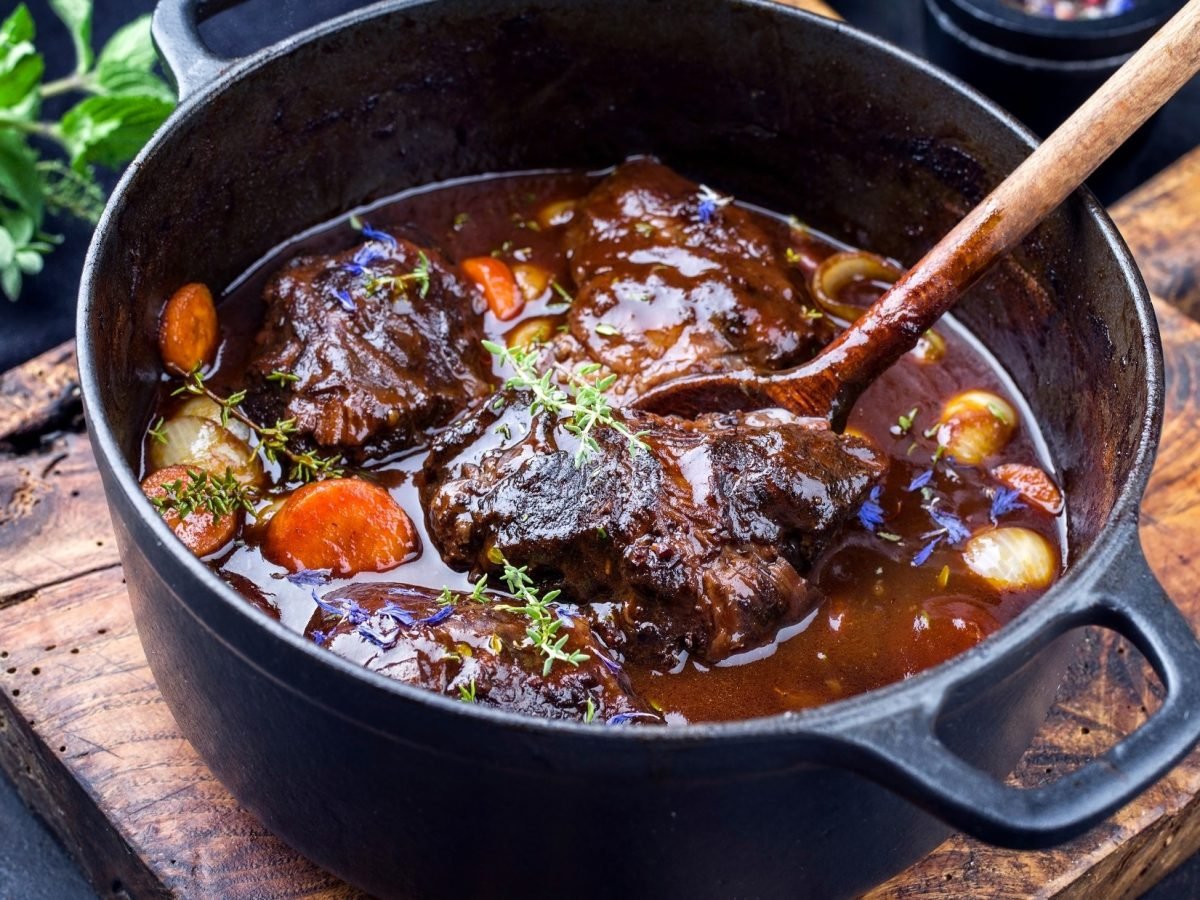 Draufsicht, schräg: Ein rustikaler Topf in dem geschmorte Rinderbäckchen in Dunkelbier-Sauce sind, steht auf einem Holzbrettchen. Und das liegt auf einem dunklen Tisch. Drumherum liegen frische Kräuter.