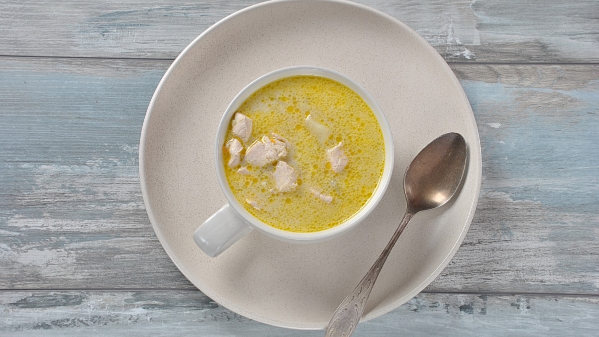 Draufsicht: Auf melierten, hellblauem Untergrund aus Holz lieget ein großer, runder und heller Teler. Auf steht eine gleichfarbige Tasse mit einer Kartoffel-Kokos-Suppe mit Hühnchen. Auf dem Teller liegt ein Suppenlöffel.