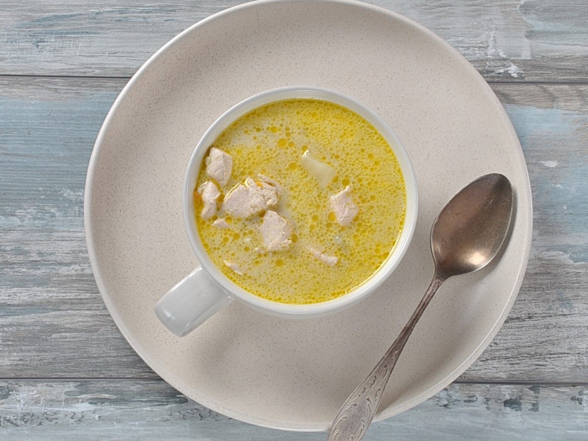 Draufsicht: Auf melierten, hellblauem Untergrund aus Holz lieget ein großer, runder und heller Teler. Auf steht eine gleichfarbige Tasse mit einer Kartoffel-Kokos-Suppe mit Hühnchen. Auf dem Teller liegt ein Suppenlöffel.