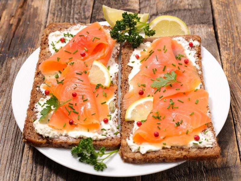 Draufsicht, schräg: zwei Lachs-Sandwich mit Hüttenkäse liegen parallel auf einem weißen Teller. Der liegt auf einem Untergrund aus Holzplanken.