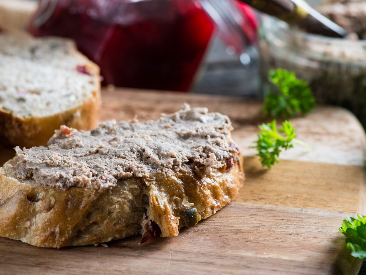 Draufsicht schräg: Auf einem Holzbrett liegt ein Weißbrot, dass mit Lamm-Leberwurst mit Masala bestrichen ist. Daneben liegen frische Petersilie und weitere Brotscheiben.