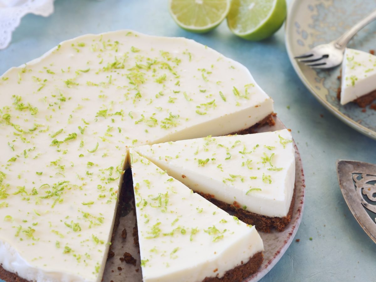 Draufsicht: Ein runder Limetten-Kuchen steht auf einem Tisch mit einer hellblauen Tischdecke. Der Kuchen ist angeschnitten. Auch auf dem Bild: zwei Kuchenstücke, ein Kuchenheber aus Metall und eine weiße Tasse mit schwarzem Kaffee.